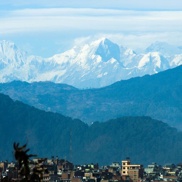 A panoramic view of a cityscape with multi-storied buildings in the foreground, lush green hills in the middle ground, and a majestic, snowy mountain range in the background under a clear blue sky.