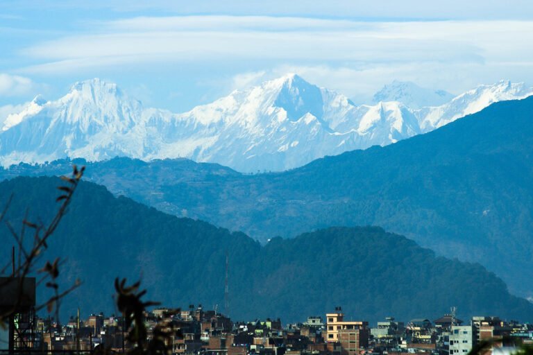 A panoramic view of a cityscape with multi-storied buildings in the foreground, lush green hills in the middle ground, and a majestic, snowy mountain range in the background under a clear blue sky.