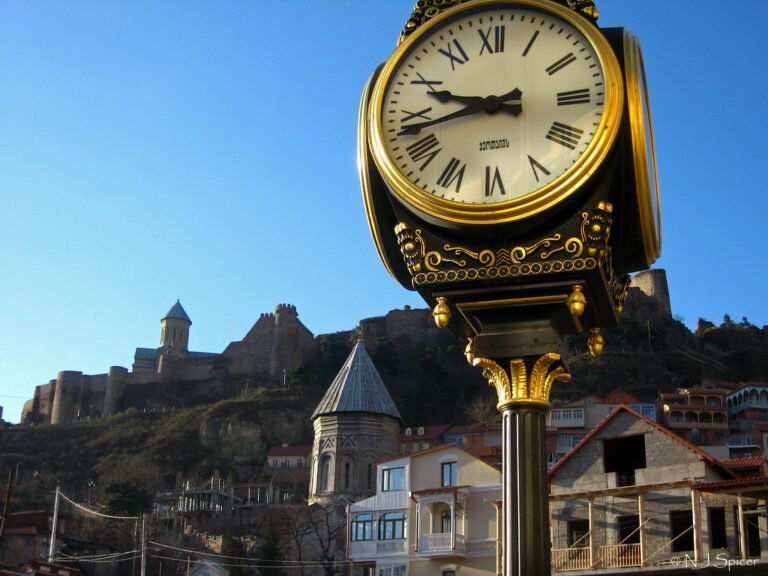 A large outdoor golden clock prominently displayed in the foreground, with an ancient fortress and buildings on a hill in the background under a clear blue sky.