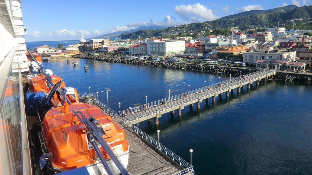 A view from a cruise ship showing a line of orange lifeboats on the side of the vessel, overlooking a long pier extending into the sea with several people walking on it, set against a coastal town backdrop with colorful buildings and green hills under a clear blue sky.