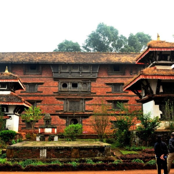 Two people walking towards an ancient multi-tiered temple with a large traditional brick building in the background, surrounded by lush greenery under a hazy sky.