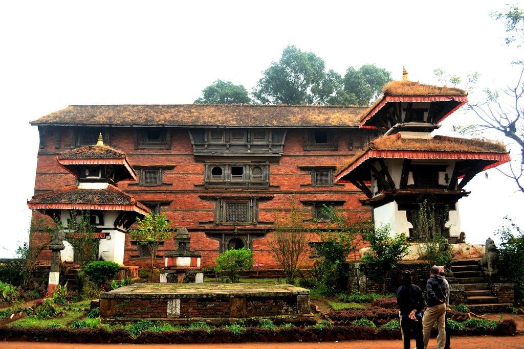 Two people walking towards an ancient multi-tiered temple with a large traditional brick building in the background, surrounded by lush greenery under a hazy sky.