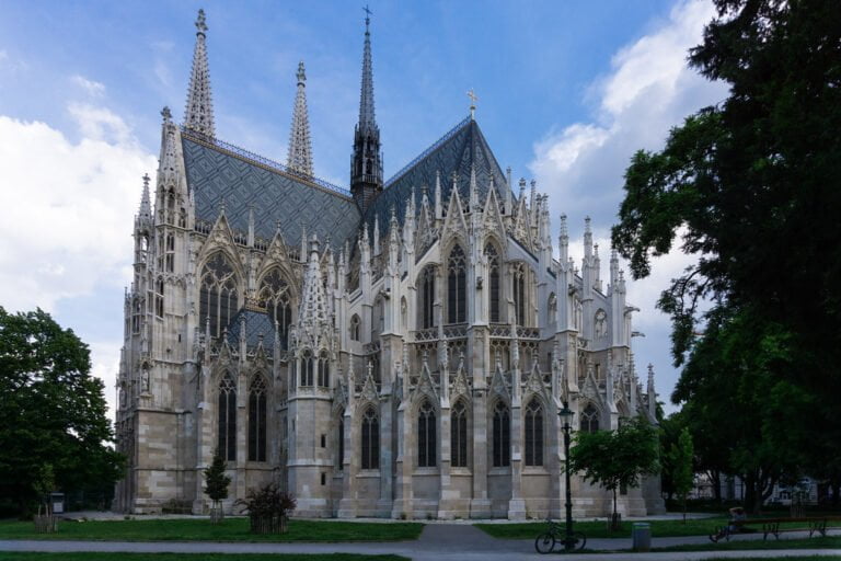 Alt text: An ornate Gothic cathedral with spired towers and intricate facade details, set against a backdrop of trees and a partly cloudy sky.