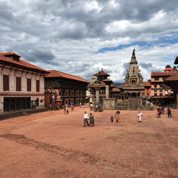 A bustling Durbar Square with traditional Nepalese architecture, featuring tiered temples, ornate palaces, open courtyards, and people walking across the brick-paved grounds under a partly cloudy sky.