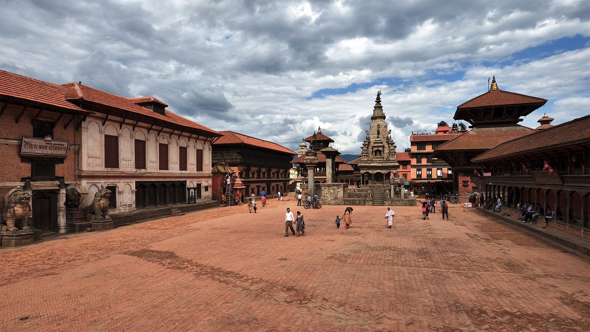 A bustling Durbar Square with traditional Nepalese architecture, featuring tiered temples, ornate palaces, open courtyards, and people walking across the brick-paved grounds under a partly cloudy sky.