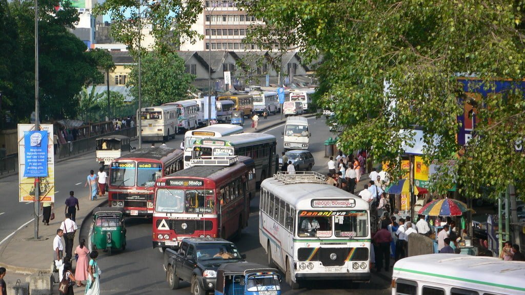 A busy street scene with multiple buses, cars, and pedestrians, some using umbrellas for shade, in an urban setting with green trees and buildings in the background.