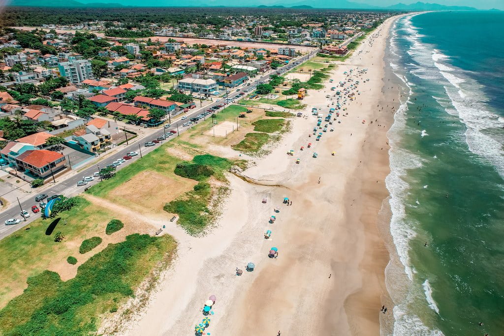 Aerial view of a bustling beach next to a coastal road with rows of houses, vehicles, and patches of greenery on a sunny day.