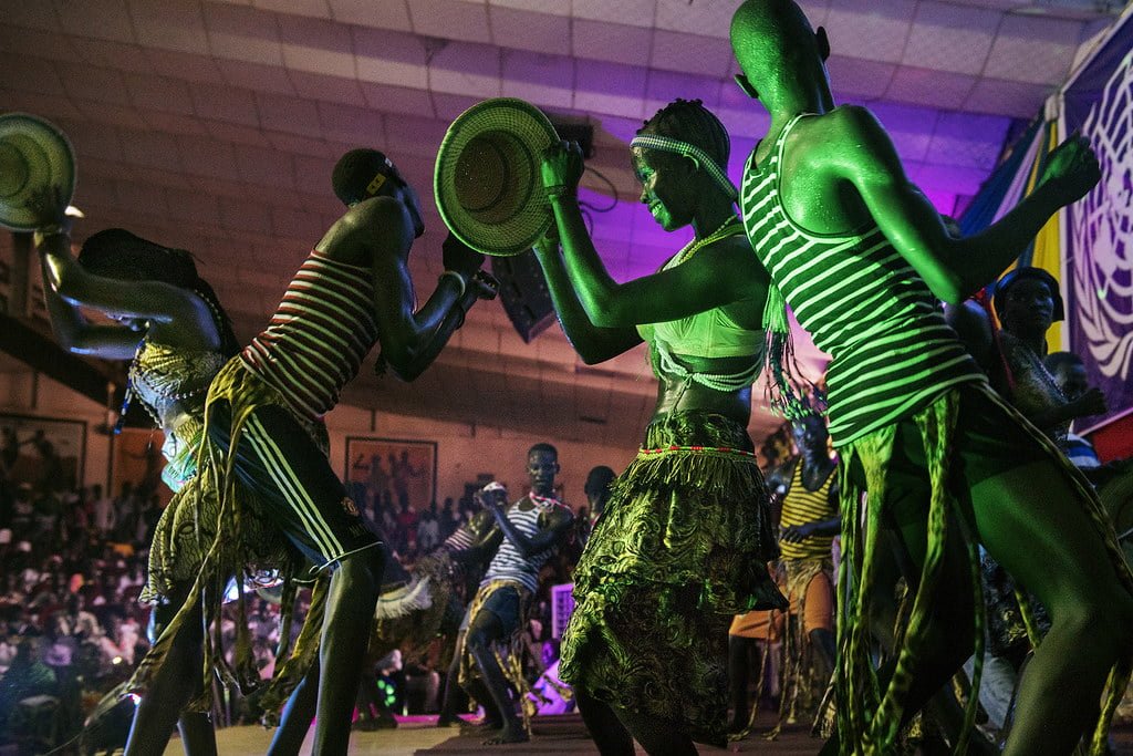 Dancers with painted bodies and traditional attire perform at a vibrant cultural event, highlighted by the intense, colorful stage lighting.