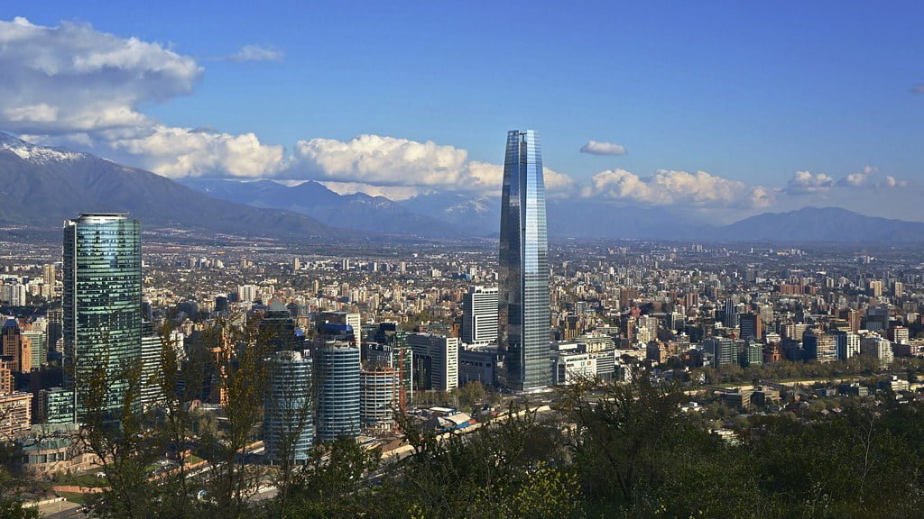 Panoramic view of a cityscape with modern skyscrapers, including a prominent glass tower, set against a backdrop of mountains under a blue sky with scattered clouds.