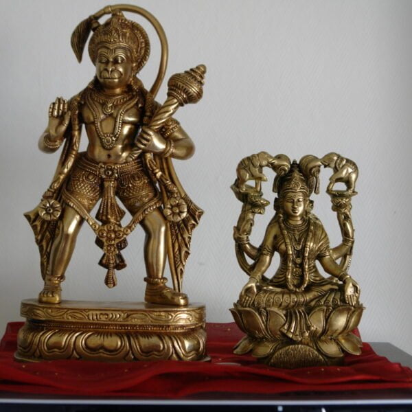 Two brass Hindu deity statues on a table with a red cloth underneath; the larger one depicts Lord Hanuman standing with a mace, and the smaller one shows Goddess Lakshmi sitting on a lotus flower.