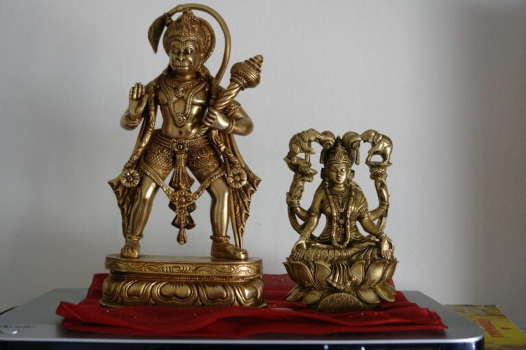 Two brass Hindu deity statues on a table with a red cloth underneath; the larger one depicts Lord Hanuman standing with a mace, and the smaller one shows Goddess Lakshmi sitting on a lotus flower.
