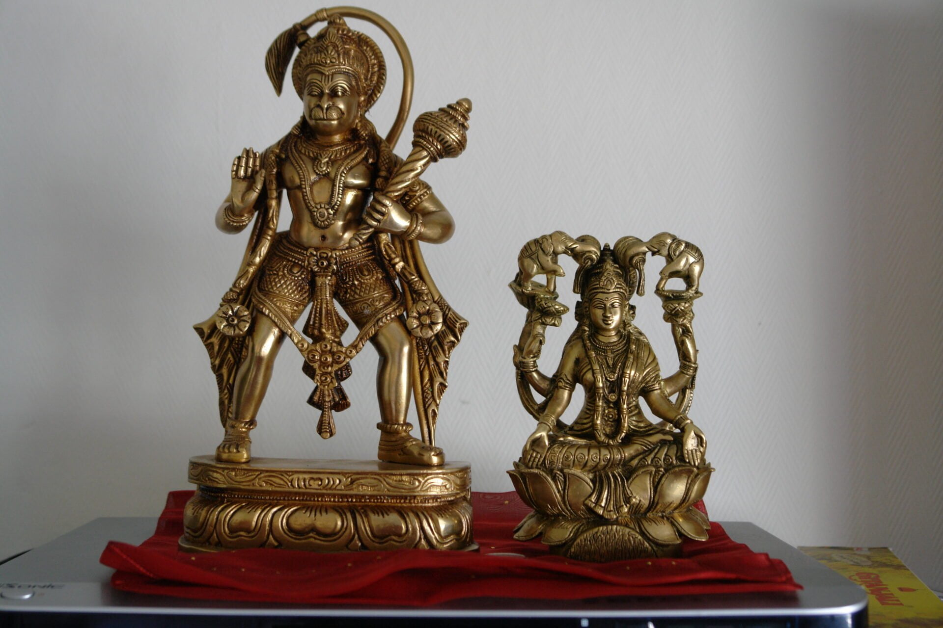 Two brass Hindu deity statues on a table with a red cloth underneath; the larger one depicts Lord Hanuman standing with a mace, and the smaller one shows Goddess Lakshmi sitting on a lotus flower.