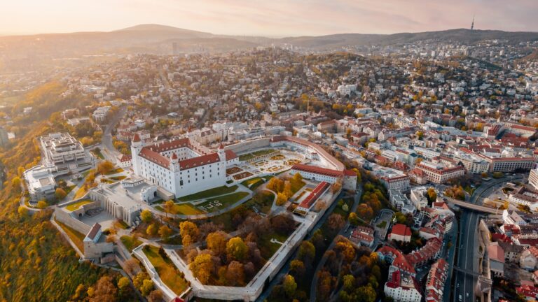Aerial view of a historic fortress complex with manicured gardens in the foreground, surrounded by modern buildings and autumn trees, with a hazy cityscape and hills in the background during sunset.