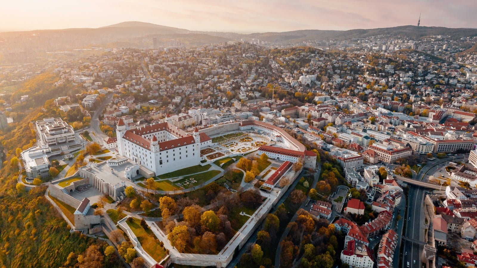 Aerial view of a historic fortress complex with manicured gardens in the foreground, surrounded by modern buildings and autumn trees, with a hazy cityscape and hills in the background during sunset.