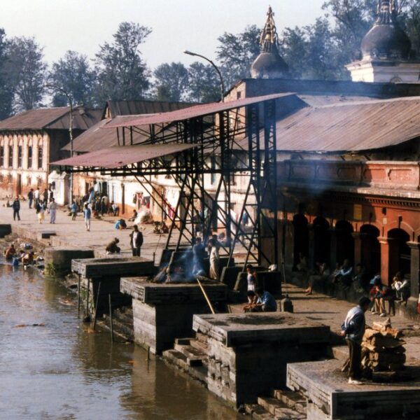 People engaging in various activities by the riverside with traditional architecture, including temples with domes, and a covered area emitting smoke.