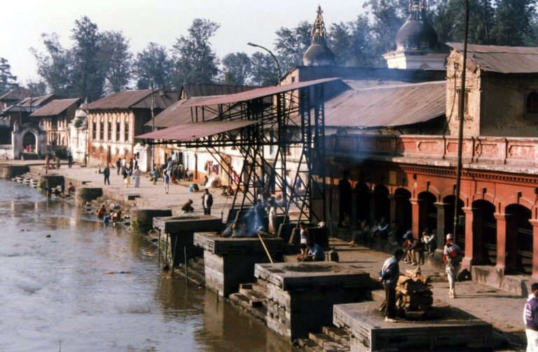 People engaging in various activities by the riverside with traditional architecture, including temples with domes, and a covered area emitting smoke.