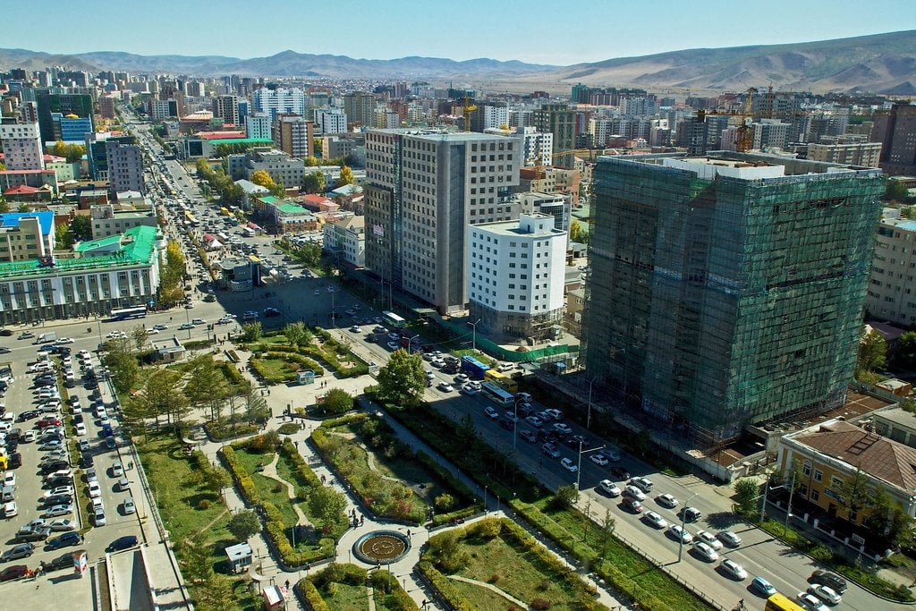 Aerial view of a bustling cityscape with various buildings, including one under construction with scaffolding, busy streets filled with traffic, and green park areas, set against a mountainous backdrop.