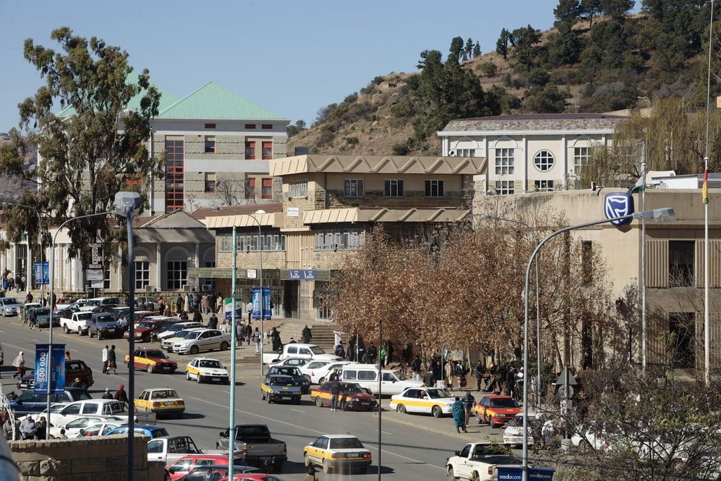 Urban street scene with busy traffic, pedestrians, and a mix of modern and traditional architecture, set against a backdrop of dry hillsides.