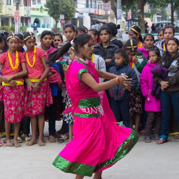 A woman in a pink and green traditional dress performing a dance on a street with a crowd of onlookers, some dressed in cultural attire.