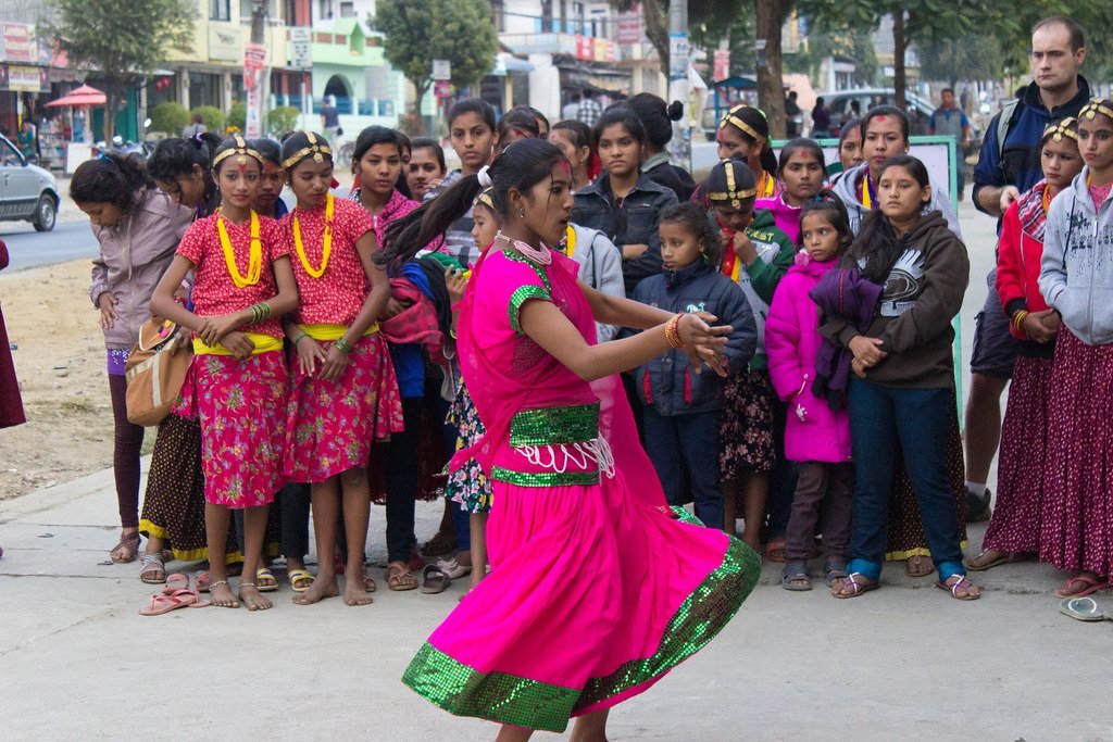 A woman in a pink and green traditional dress performing a dance on a street with a crowd of onlookers, some dressed in cultural attire.
