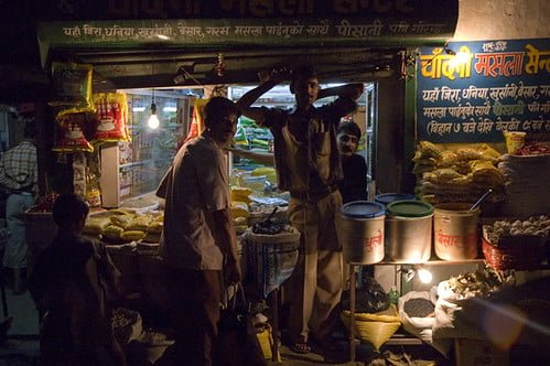 Street vendors at a dimly lit market stall in India at night, displaying various food items, with signage in Hindi above them.