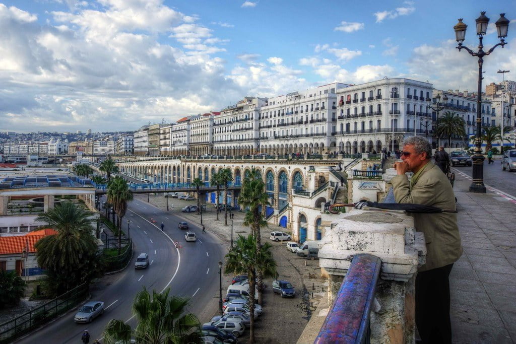 A man looking over a bustling city street lined with white buildings and arches, palm trees, and a partly cloudy sky above.