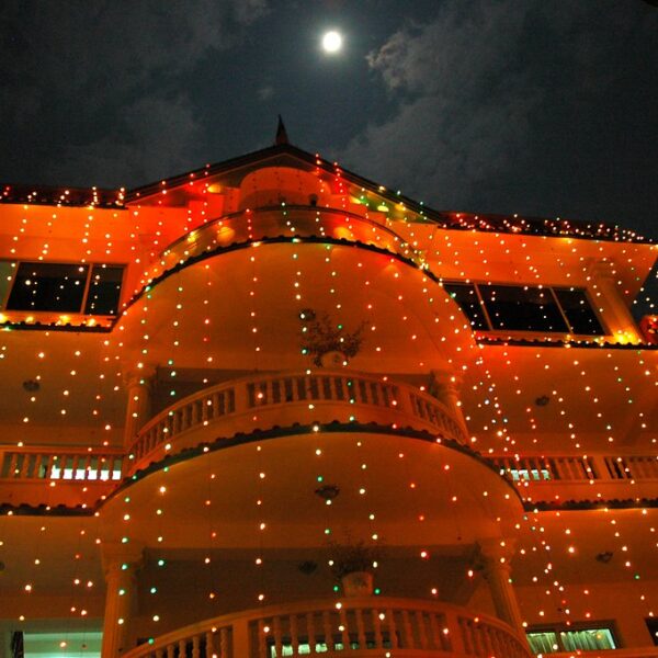 A large multi-storied building at night adorned with colorful string lights, with the moon partially visible in the sky above.