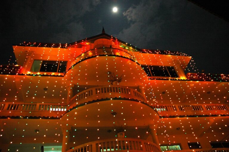 A large multi-storied building at night adorned with colorful string lights, with the moon partially visible in the sky above.