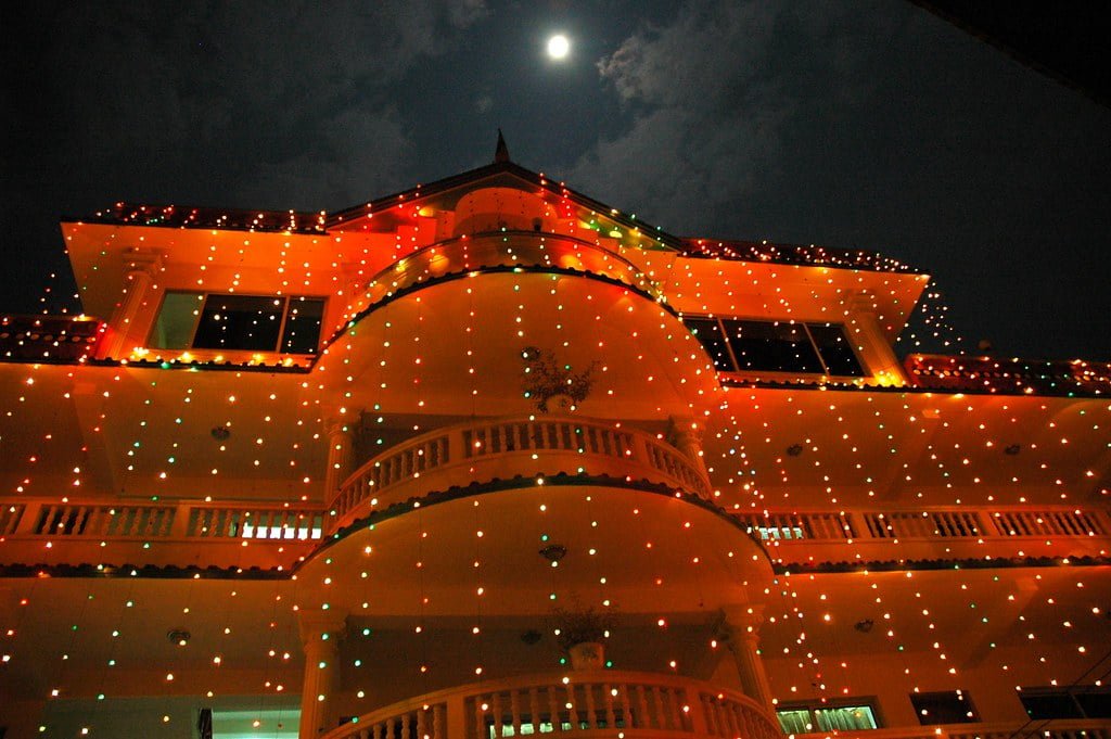 A large multi-storied building at night adorned with colorful string lights, with the moon partially visible in the sky above.