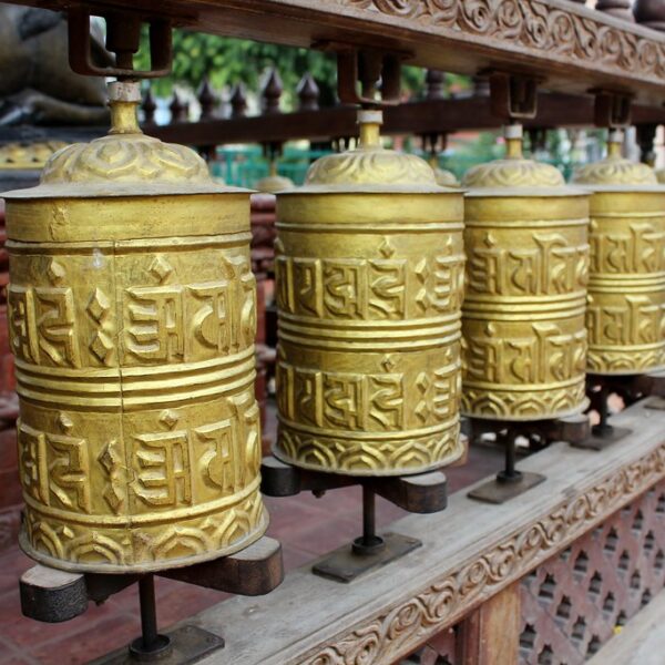 A row of brass Tibetan prayer wheels with embossed mantras on a wooden stand.