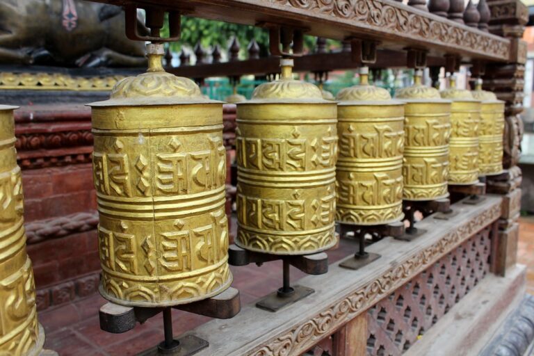 A row of brass Tibetan prayer wheels with embossed mantras on a wooden stand.