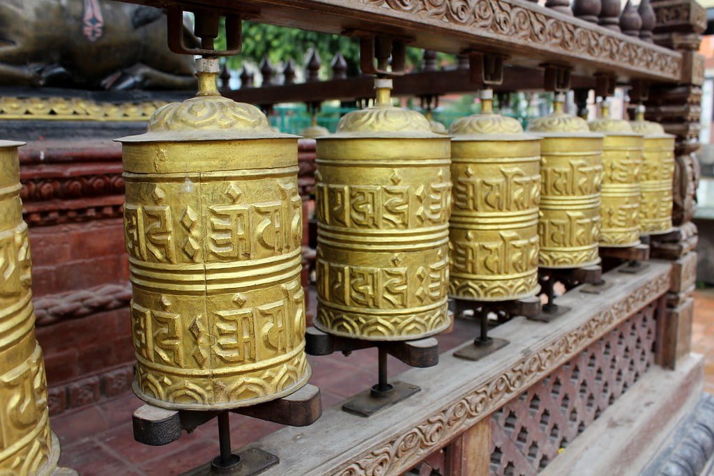 A row of brass Tibetan prayer wheels with embossed mantras on a wooden stand.
