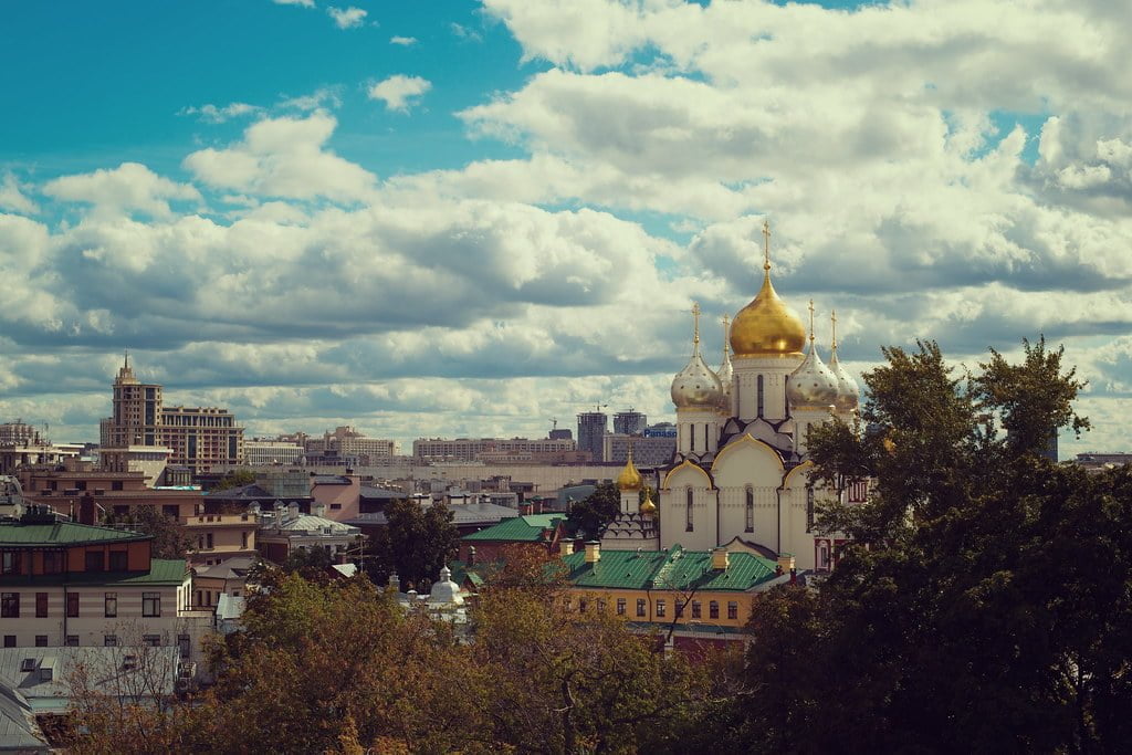 A cityscape with a mix of historical and modern buildings under a cloudy sky, featuring a prominent church with golden and silver domes.