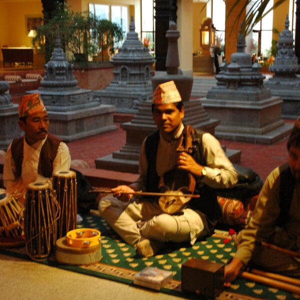 Three musicians in traditional Nepalese attire performing inside a hall with cultural architecture and decor, playing the tabla, sarangi, and flute.