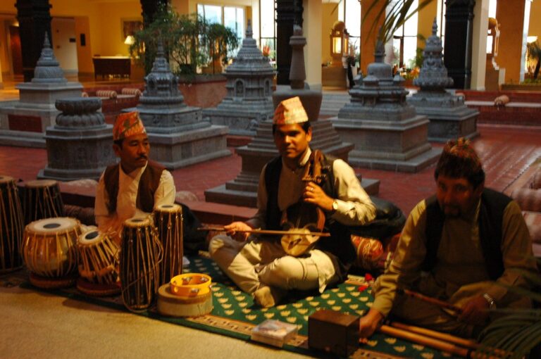 Three musicians in traditional Nepalese attire performing inside a hall with cultural architecture and decor, playing the tabla, sarangi, and flute.