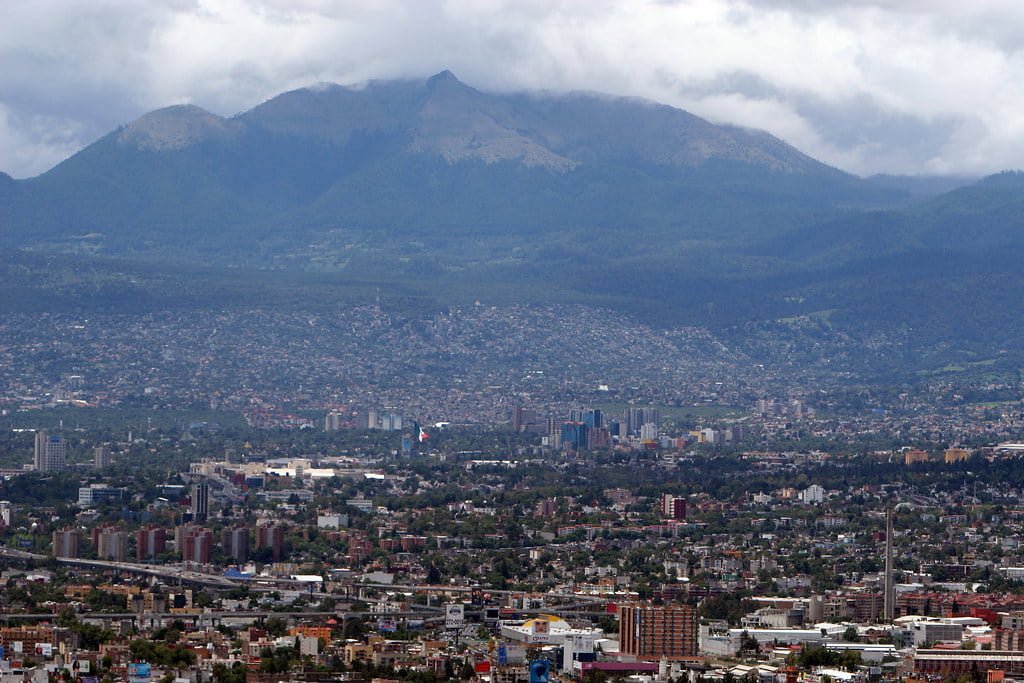 A panoramic view of a sprawling city with numerous buildings in the foreground leading up to tree-covered mountains shrouded in clouds in the background.