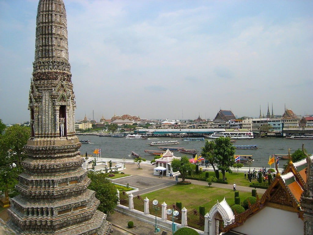 View from Wat Arun featuring a tall, intricately decorated spire with the Chao Phraya River and Bangkok skyline in the background.