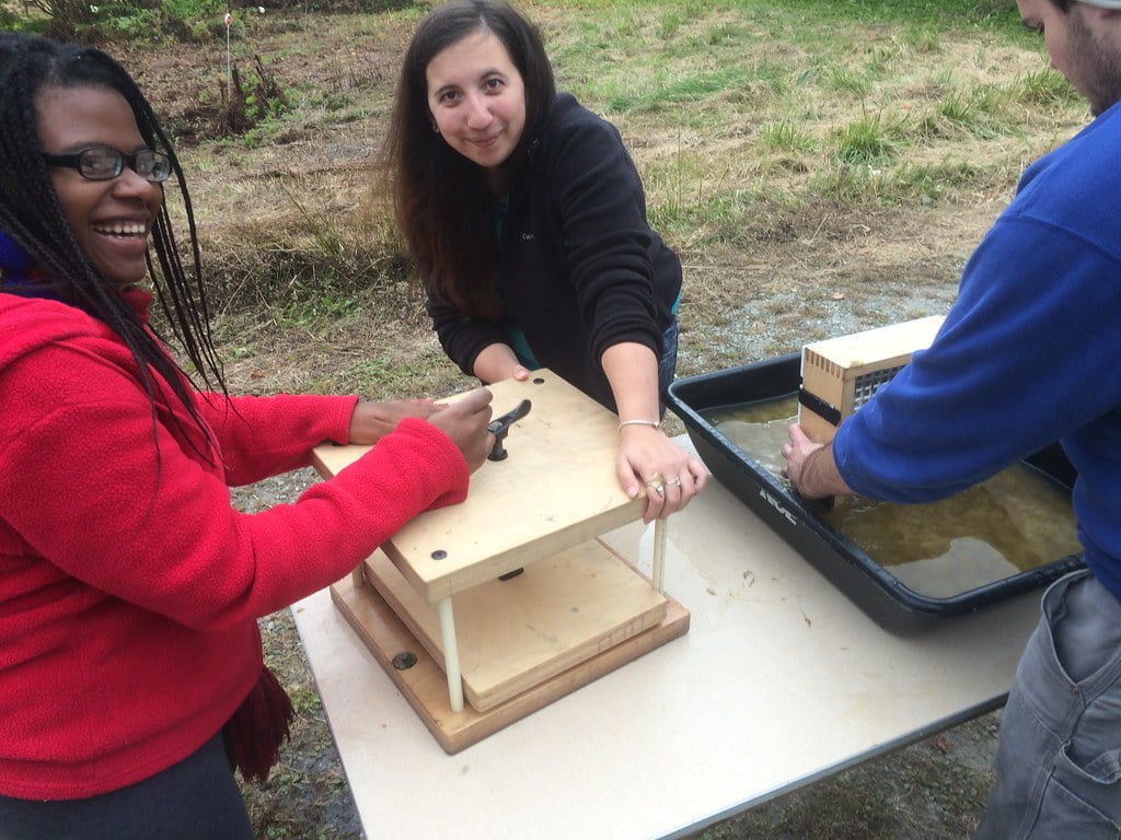 Three people around a table engaged in a hands-on experiment or educational activity outdoors, using a wooden apparatus and a tray of water.