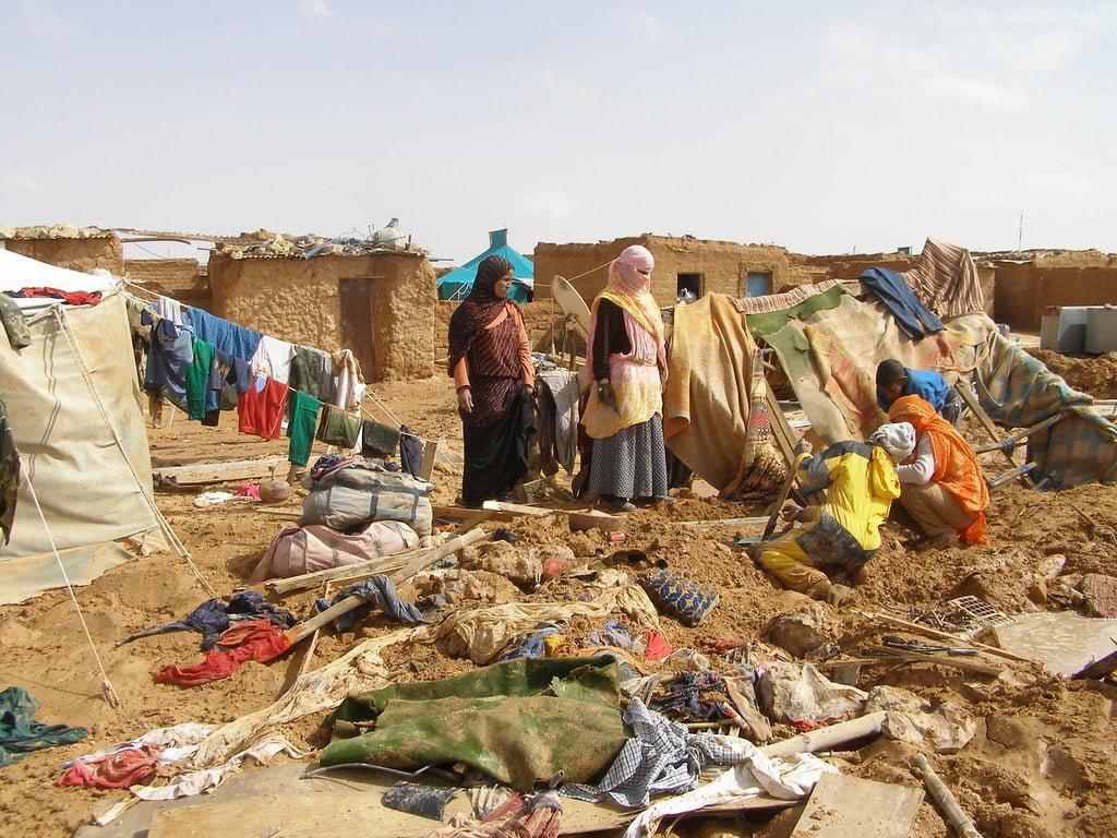 A group of people in a makeshift settlement with mud houses and debris, with some individuals standing and one person squatting while handling materials on the ground. Clothes are hung out to dry on a line, and the ground appears muddy and littered with scattered belongings.