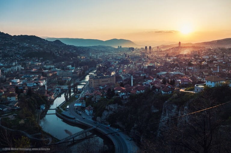A panoramic view of a city at sunset with sunlight casting a warm glow over buildings, a river cutting through the center, and surrounding hills under a soft sky.