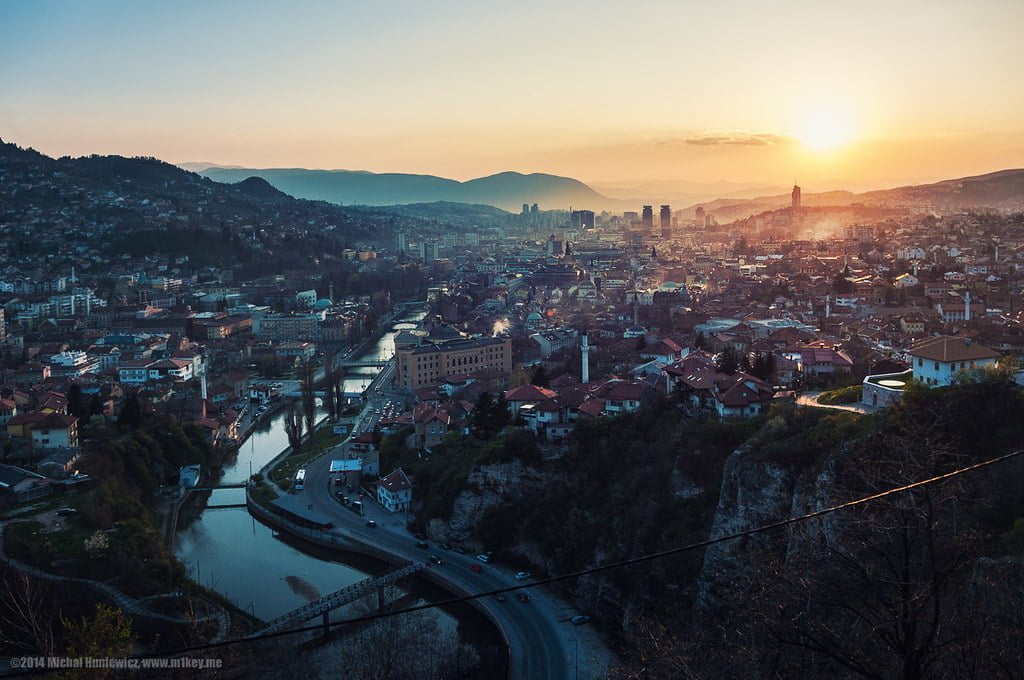 A panoramic view of a city at sunset with sunlight casting a warm glow over buildings, a river cutting through the center, and surrounding hills under a soft sky.