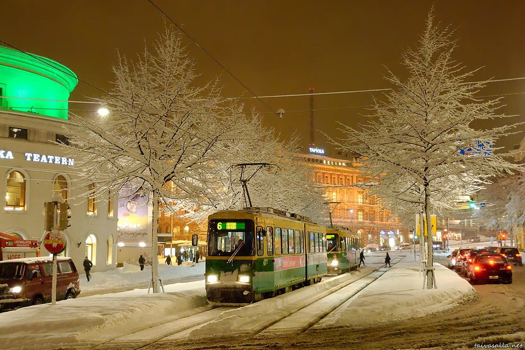 Tram on snowy tracks at an urban street at night with illuminated buildings and trees covered in snow.