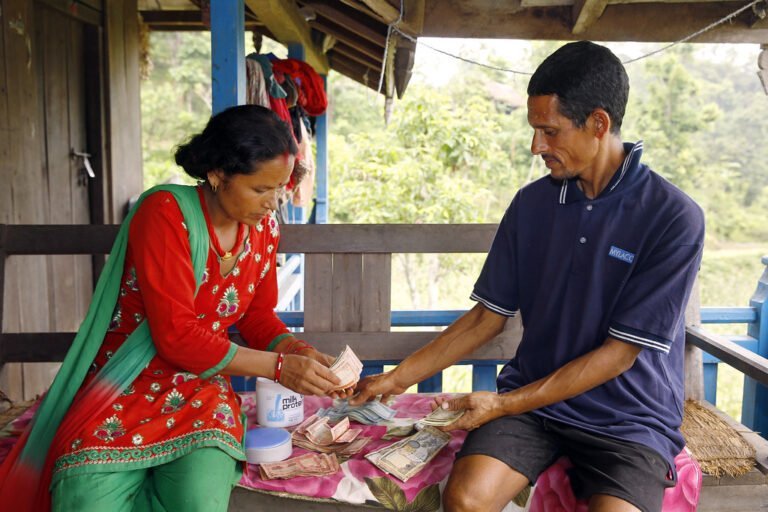 A woman in a red and green sari counts money while sitting across from a man in a blue striped polo shirt who is also handling cash, both seated on a porch with a rural background.