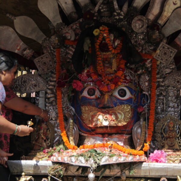 A person making an offering at an elaborately decorated shrine with a large, colorful mask surrounded by flowers and traditional ornaments.