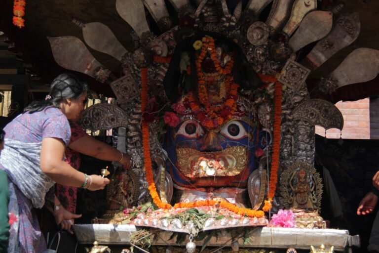 A person making an offering at an elaborately decorated shrine with a large, colorful mask surrounded by flowers and traditional ornaments.