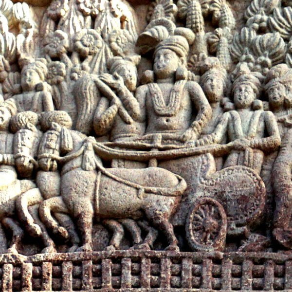 Ancient stone relief depicting a chariot pulled by horses, surrounded by various figures, some appearing to be in conversation, and intricate foliage patterns above.