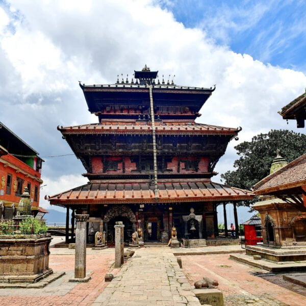 Alt text: An ornate multi-tiered temple with elaborate wood carvings and brickwork, standing in a courtyard surrounded by brick structures with traditional Nepalese architectural elements under a partly cloudy sky.