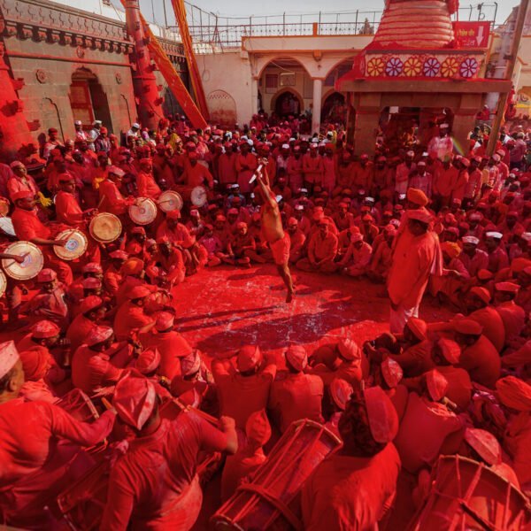 A vibrant scene with numerous people dressed in red gathered around a central figure who appears to be throwing a cloud of red powder, with others playing drums and celebrating amidst a red-soaked environment, indicating a festive or ceremonial event.