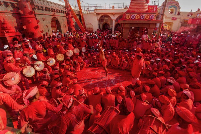 A vibrant scene with numerous people dressed in red gathered around a central figure who appears to be throwing a cloud of red powder, with others playing drums and celebrating amidst a red-soaked environment, indicating a festive or ceremonial event.