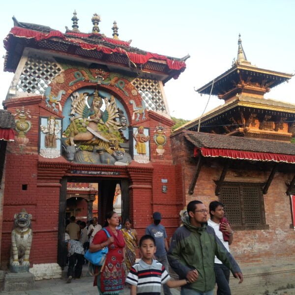 Alt text: A vibrant entrance of a traditional Nepalese temple adorned with multi-colored wood carvings of a deity surrounded by mythical creatures, flanked by stone lion statues, with people walking by and a two-tiered pagoda-style roof visible in the background.